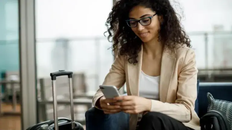 Mujer rizada y con gafas interactúa con su celular al lado de su equipaje en sala de espera de aeropuerto